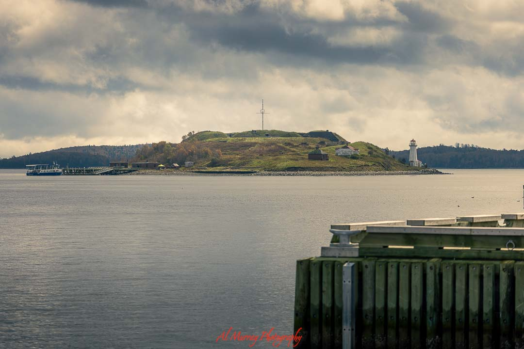 Wide landscape photo of a small island with a white lighthouse, grassy hill, and scattered buildings surrounded by calm harbor waters under a cloudy sky. Viewed from a wooden pier in the foreground, the peaceful coastal scene reflects a scenic stop on a Maritimes driving tour for seniors from Halifax, showcasing accessible waterfront views and classic Atlantic charm.
