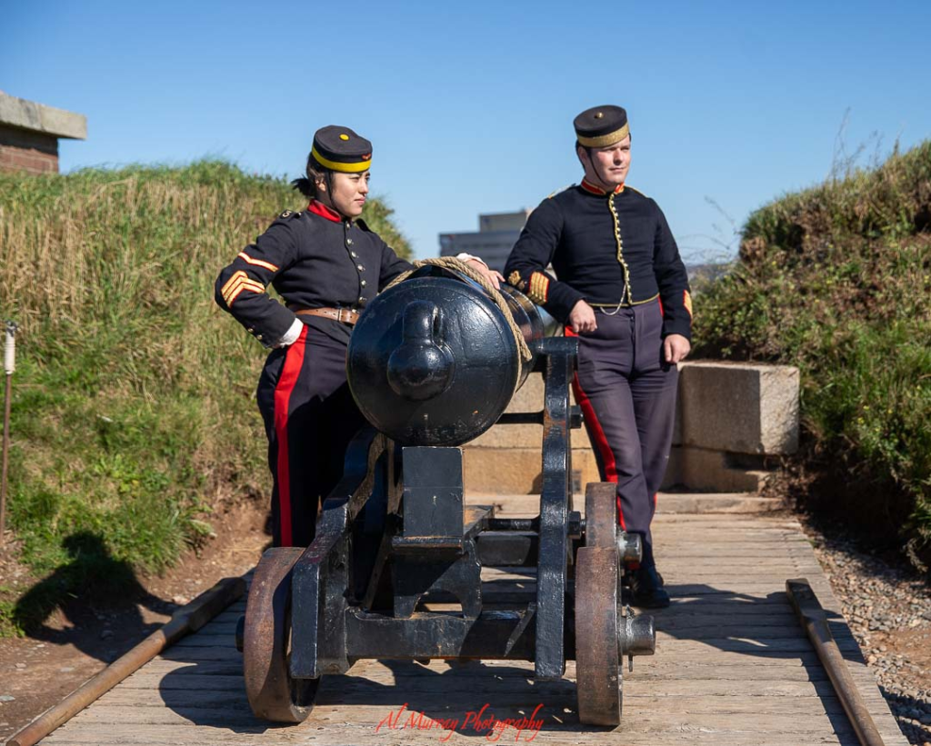 Two historical interpreters in dark military uniforms stand beside a large black cannon mounted on wooden wheels at Citadel Hill. The grassy earthworks and wooden walkway highlight the historic fort setting, capturing the living history experience visitors can explore at Citadel Hill in Halifax.
