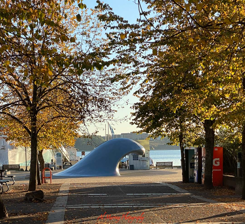 Photo of a large blue wave sculpture on a waterfront plaza framed by trees with golden autumn leaves and calm harbor water in the background. The peaceful boardwalk setting captures a scenic stop on a Bay of Fundy road trip, highlighting coastal art and fall colors along the Maritime shoreline.