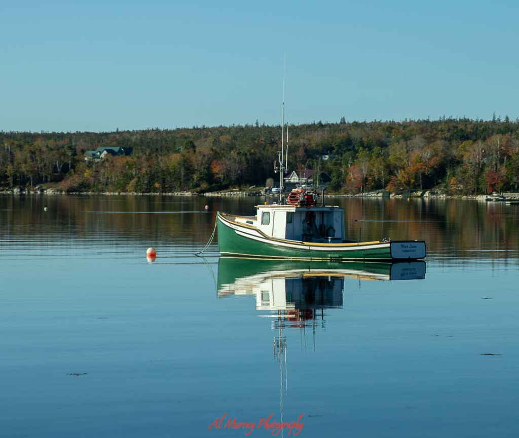 Photo of a small green and white fishing boat floating on calm glassy water with its reflection mirrored below and a tree lined shoreline in the distance. The peaceful harbor scene captures the quiet maritime beauty travelers can enjoy on a New Brunswick coastal drive, highlighting charming seaside communities and working waterfronts.
