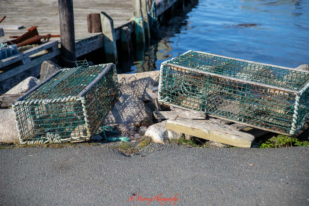 Photo of two green wire lobster traps resting on a wooden dock beside calm blue harbor water. The working waterfront scene highlights the fishing heritage and coastal charm travelers can experience on a PEI road trip from Halifax, with fresh seafood and maritime culture along the way.