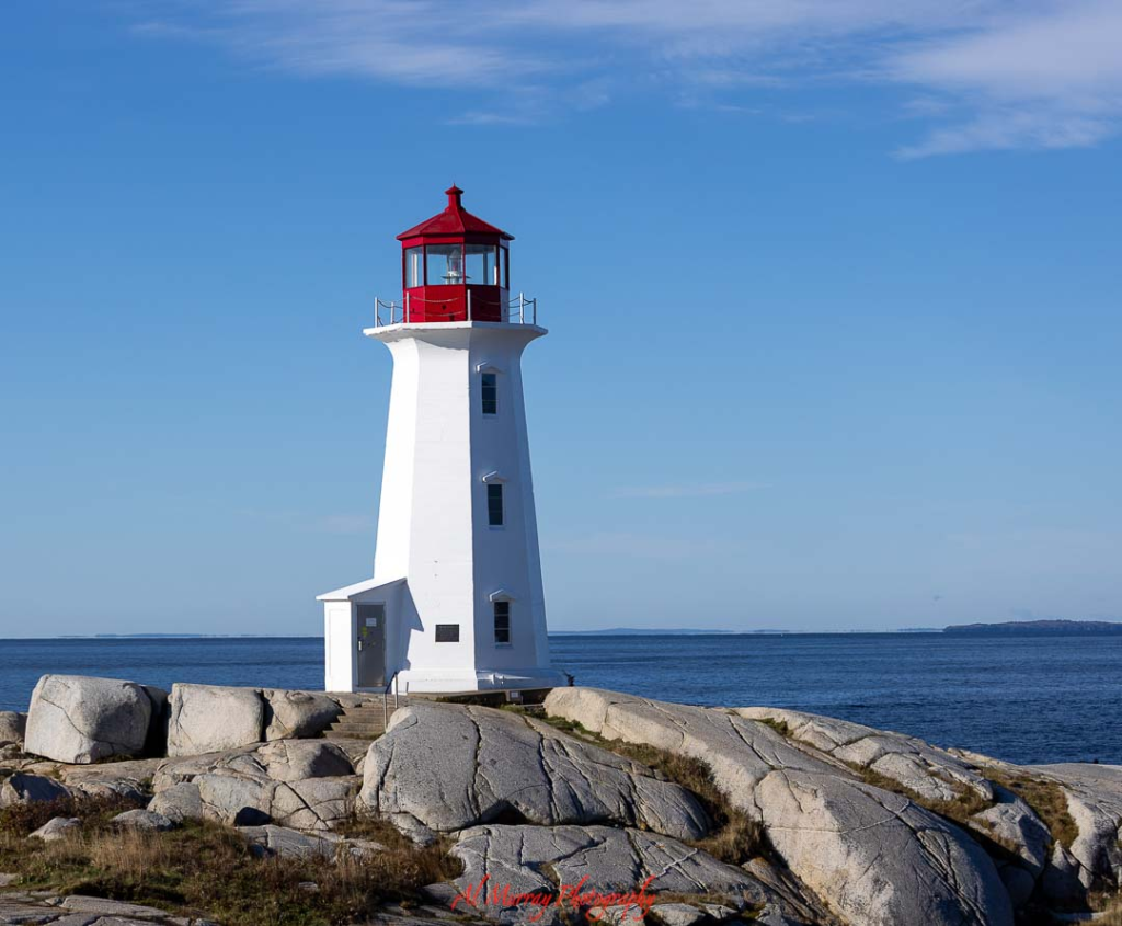 Photo of a white lighthouse with a red lantern room perched on smooth granite rocks overlooking the Atlantic Ocean under a clear blue sky. The iconic coastal landmark captures the highlight of a Peggy’s Cove day trip from Halifax, showcasing the rugged shoreline and classic Nova Scotia scenery visitors come to see.