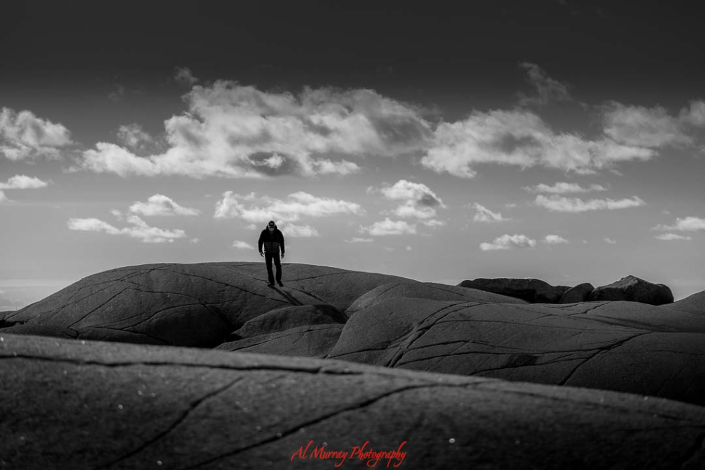 Black and white photo of a senior man walking alone across large smooth coastal rock formations under a wide sky filled with scattered clouds. The dramatic landscape and open horizon evoke a peaceful stop along a Maritimes driving tour for seniors from Halifax, highlighting the quiet beauty and accessible seaside scenery of Atlantic Canada.