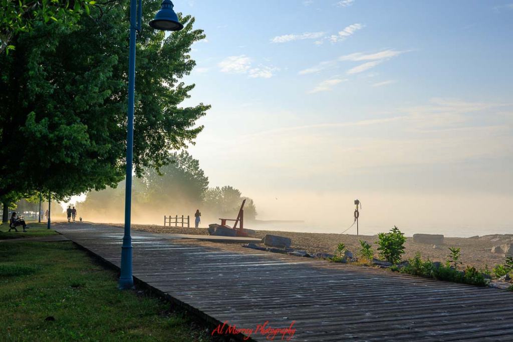 where to watch the world cup 2026 in Toronto Wooden boardwalk along a sandy Toronto beach at sunrise with soft mist over the water, trees casting shade on the path, and a few people walking in the distance. The peaceful lakeside scene captures outdoor Things to do in Toronto during World Cup 2026, offering a relaxing break between matches.