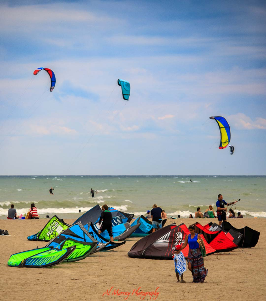 Colorful kites and kiteboards scattered across a sandy beach while surfers ride waves under bright blue skies, capturing an energetic waterfront scene in Toronto. The mix of spectators relaxing on the shore and active water sports highlights senior friendly things to do in Toronto, from scenic beach walks to enjoying lively lakeside views and outdoor recreation.