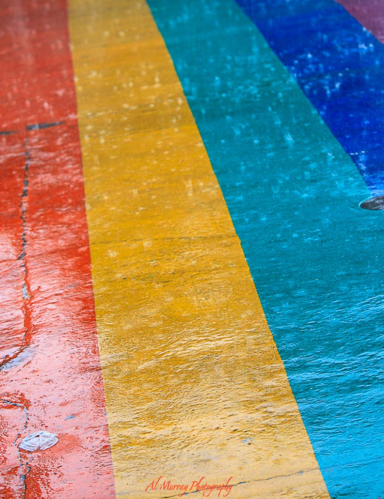 Close up of a rainbow painted sidewalk with bold red orange yellow green blue and purple stripes glistening with rain, symbolizing pride and inclusion in the city. The vibrant colors and wet pavement capture the welcoming spirit of LGBTQ friendly Toronto travel and highlight a supportive urban community atmosphere.