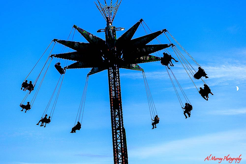 Silhouette of a tall swing ride spinning against a bright blue sky with riders suspended in the air and a faint moon visible in the background, capturing the lively atmosphere of Toronto summer festivals for adults over 50. The dramatic upward view and moving chairs highlight the excitement and outdoor entertainment that make seasonal city events memorable and engaging.
