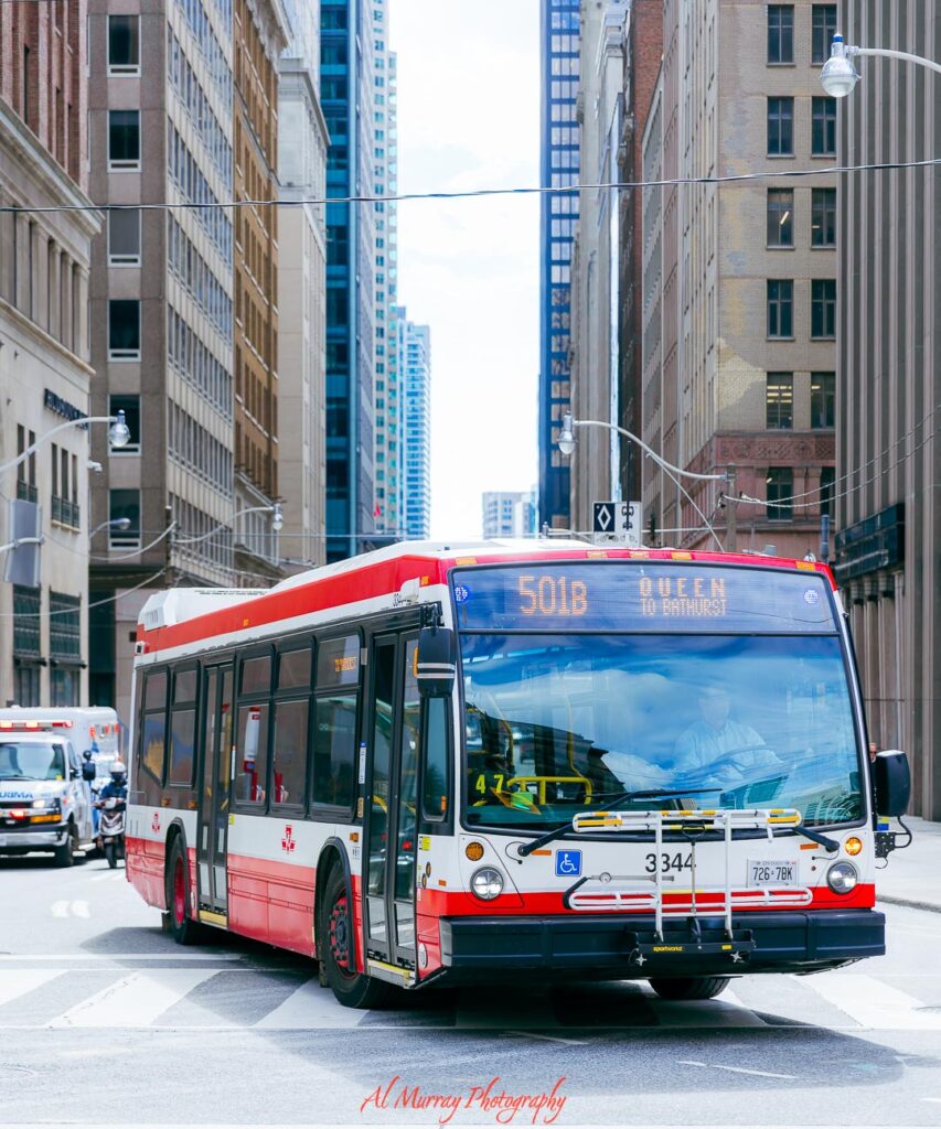 Red and white city bus labeled "501B Queen to Bathurst" driving through a downtown Toronto street lined with tall buildings, illustrating how to get around areas highlighted in where to stay in Toronto without a car.