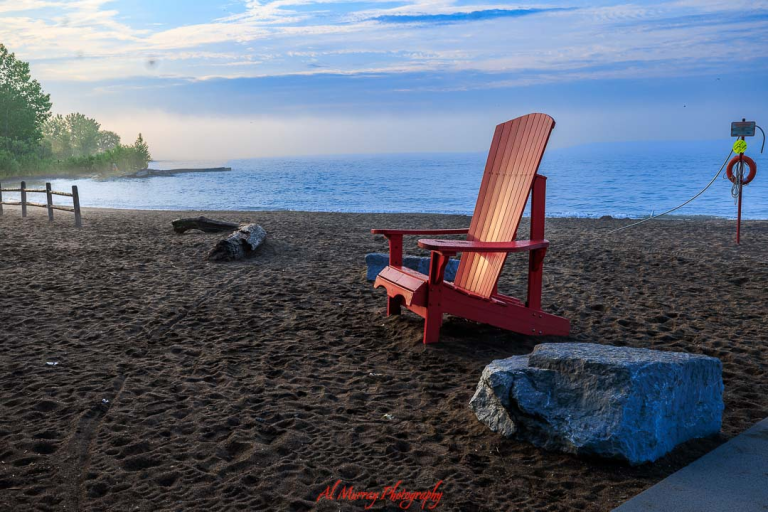Red Adirondack chair facing Lake Ontario on a quiet sandy beach at sunrise with soft blue sky and gentle waves, capturing a peaceful lakeside scene ideal for toronto spring and summer travel for seniors. Large rocks, a fallen log, and a lifebuoy post add detail to the shoreline, highlighting a calm and accessible waterfront setting perfect for relaxing seasonal getaways.
