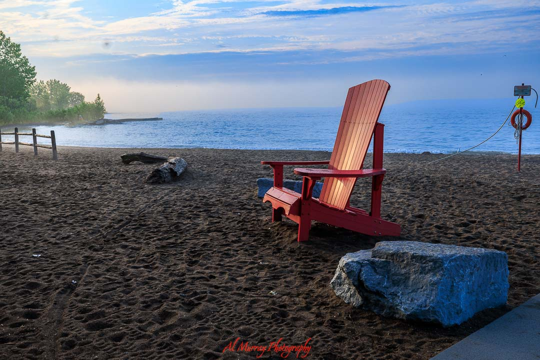 Red Adirondack chair facing Lake Ontario on a quiet sandy beach at sunrise with soft blue sky and gentle waves, capturing a peaceful lakeside scene ideal for toronto spring and summer travel for seniors. Large rocks, a fallen log, and a lifebuoy post add detail to the shoreline, highlighting a calm and accessible waterfront setting perfect for relaxing seasonal getaways.