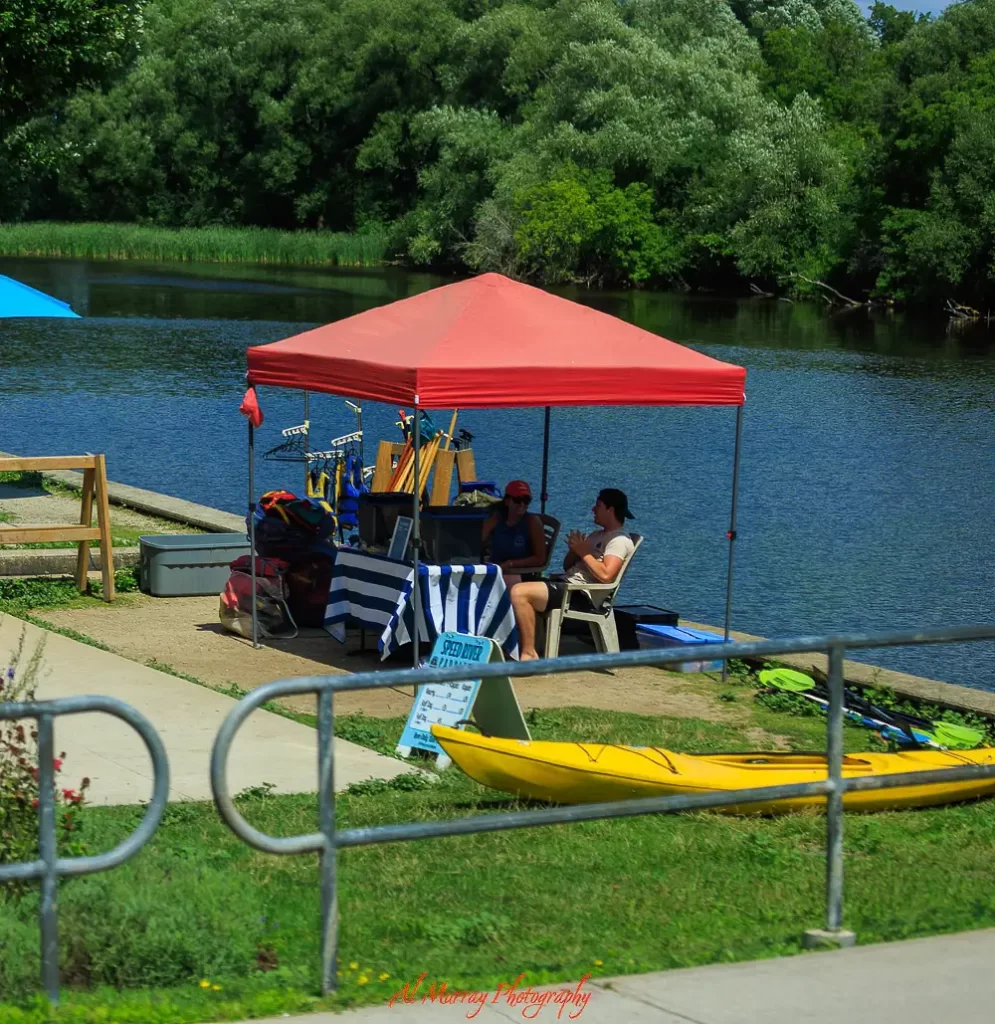 Two people sit under a red canopy beside a calm river offering kayak rentals, with paddles and a bright yellow kayak nearby, showing a relaxed outdoor activity for spring and summer getaways from Toronto for seniors.
