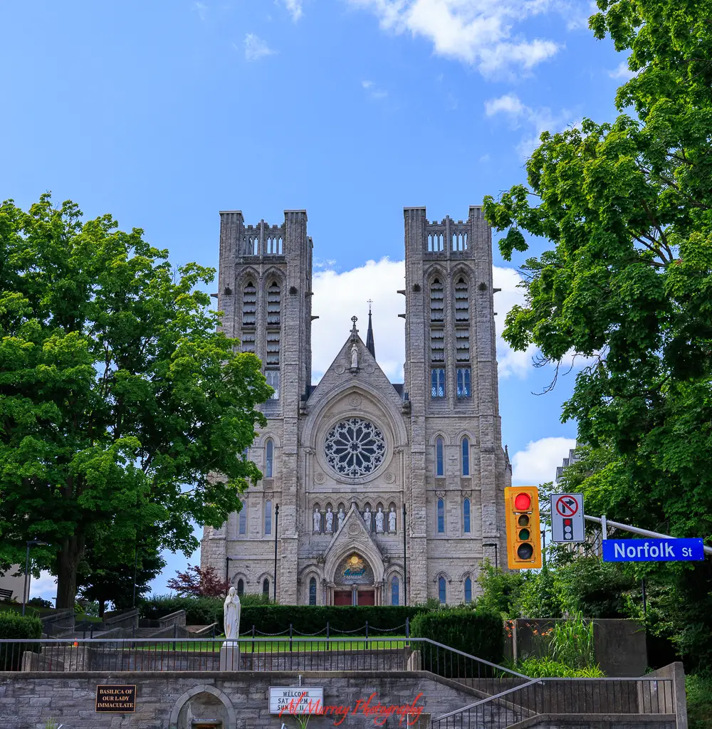 Large stone cathedral with twin towers and a circular stained glass window stands at a street corner with a visible Norfolk Street sign, highlighting a historic landmark that can be visited on cheap day trips from Toronto by train.