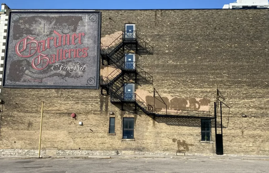 Brick building with a large painted sign reading "Estate Gardner Galleries Fine Art" and a black metal fire escape zigzagging up the wall, reflecting a charming small town stop along scenic drives from Toronto Ontario.