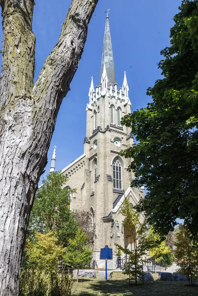 Tall stone church with a pointed steeple rises above leafy trees on a sunny day, highlighting a historic small town stop ideal for easy getaways from Toronto without a car.