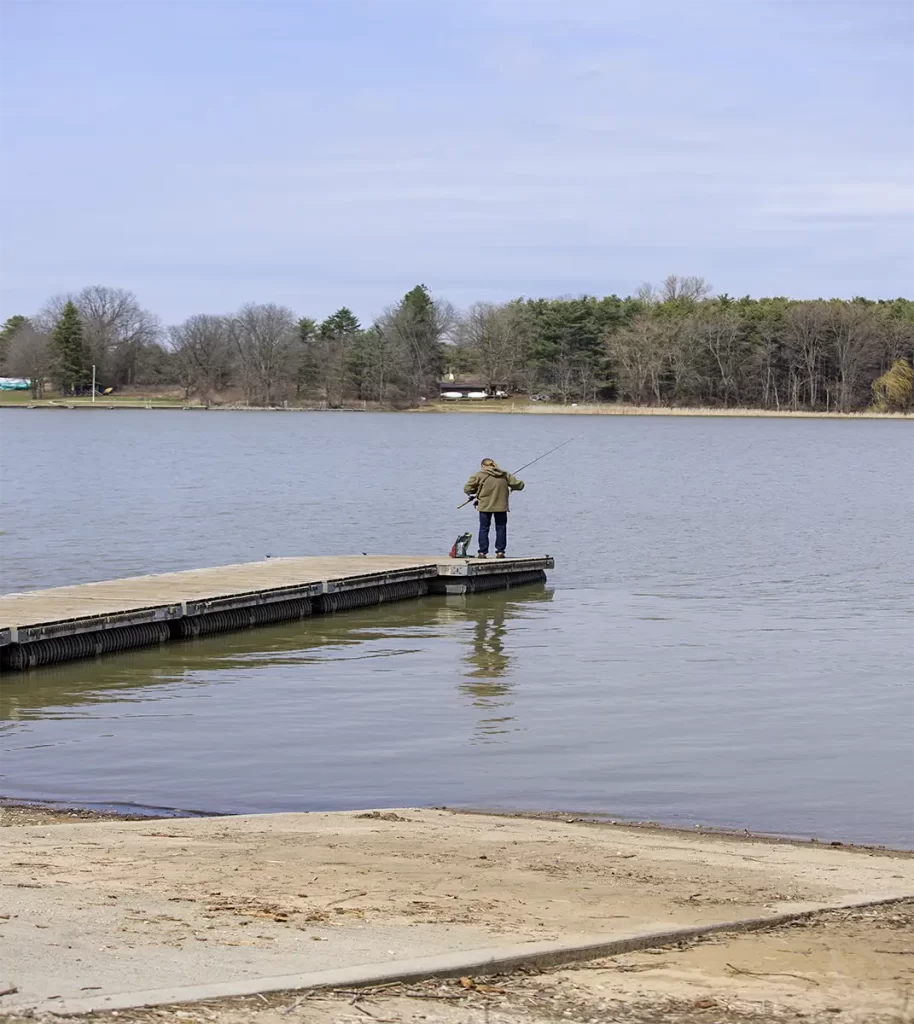 Person standing at the end of a wooden dock fishing on a calm lake with trees lining the opposite shore, capturing a quiet and accessible outdoor activity for day trips from Toronto by train.