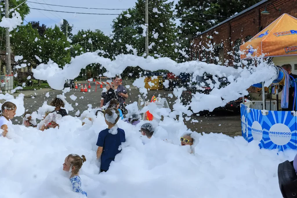 Children play in a street filled with foam as bubbles float through the air beside a small event tent labeled "Presswood Entertainment," capturing a fun local festival atmosphere for short trips from Toronto Ontario.