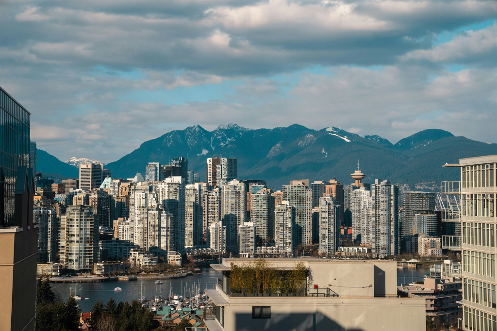 Vancouver skyline with dense high rise buildings along the waterfront and snow capped mountains rising behind the city under a partly cloudy sky. The dramatic mix of ocean city and mountains highlights why Vancouver is featured in many British Columbia summer travel ideas.