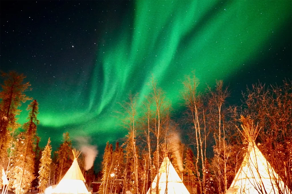 Bright green northern lights sweeping across a star filled night sky above illuminated teepees surrounded by tall trees, creating a dramatic winter wilderness scene. The glowing aurora and cozy camp setting capture the magic of Yellowknife northern lights tours, highlighting guided nighttime experiences in Canada’s far north.