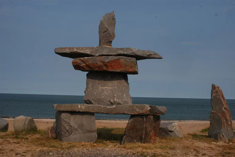 Inukshuk stone sculpture stacked with large flat rocks standing on a quiet shoreline with calm blue water and open sky in the background, representing a northern cultural landmark. The balanced stones and remote coastal setting capture the spirit of Canadian North Travel for Seniors, highlighting meaningful heritage sites and peaceful scenic landscapes ideal for reflective exploration.