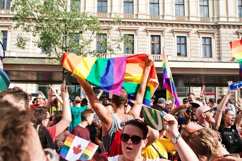 Crowd celebrating at a Pride parade while a person holds a large rainbow flag above their head and others wave pride and Canadian flags. The festive street scene reflects the vibrant atmosphere of Canada Pride festivals held in cities across the country. 🌈