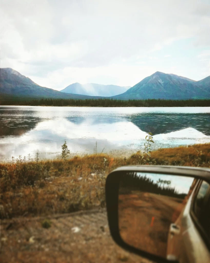 View from a vehicle parked beside a calm northern lake with mountain peaks reflected in the water and soft sunlight breaking through clouds in the distance. The open wilderness landscape and quiet roadside perspective capture the scenic freedom of Canadian North travel for seniors, highlighting peaceful drives and accessible natural viewpoints.
