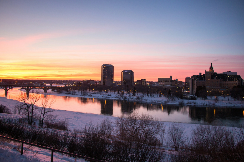 City skyline at sunset reflected in a calm river with a historic hotel and bridge silhouetted against pink and orange winter sky. The glowing evening view shows a scenic prairie city stop along a road trip western canada itinerary.