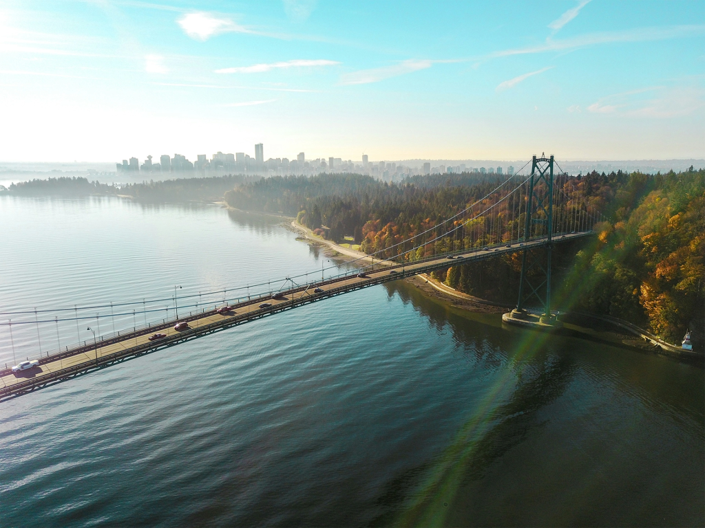 Aerial view of a long suspension bridge crossing calm ocean water with forested shoreline and a distant city skyline under a bright blue sky. The scenic coastal landscape showcases the type of views visitors experience during spring and summer travel in British Columbia.