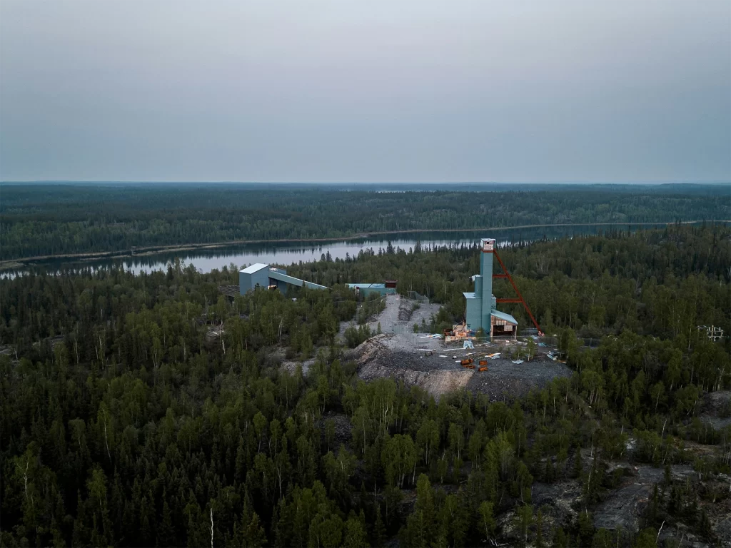 Aerial view of a remote northern landscape with dense green forest surrounding a small industrial site near a winding river under an overcast sky. The vast wilderness setting reflects canadian north travel for seniors, highlighting guided excursions to unique northern communities and the expansive natural scenery of Canada’s far north.