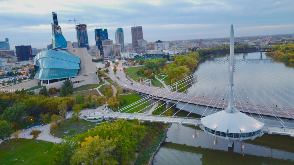 Aerial view of a modern cable stayed pedestrian bridge crossing a wide river beside a striking glass museum building and downtown skyline. The city scene highlights an urban stop travelers might explore on a road trip western canada route.