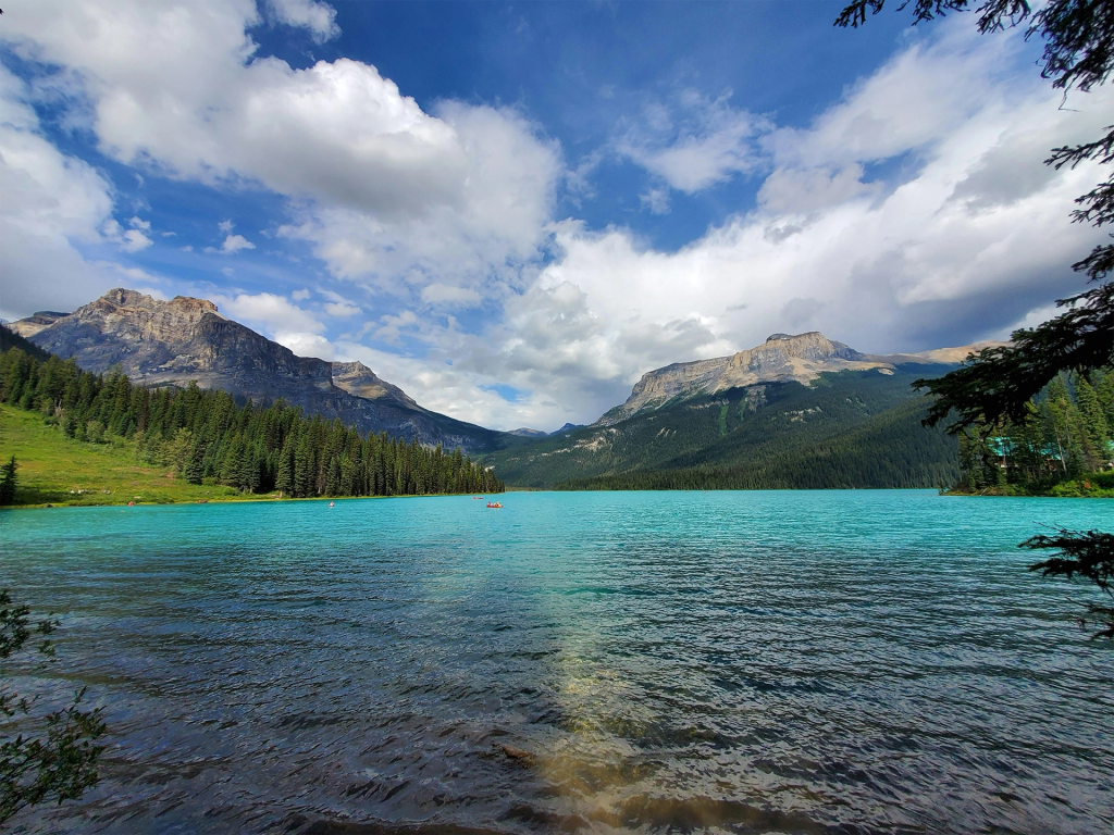 Turquoise glacial lake surrounded by evergreen forest and rugged mountains beneath a wide sky filled with dramatic white clouds. Tiny red canoes dot the water highlighting the outdoor adventures popular during spring and summer travel in western canada.