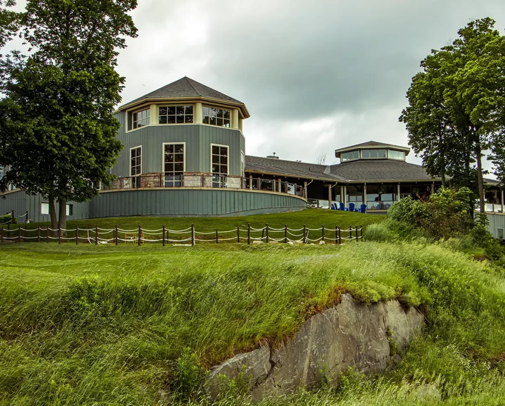Large lakeside building with wraparound windows and a covered patio sits on a grassy hill overlooking the water, suggesting a scenic stop along one of the best road trips from Toronto.