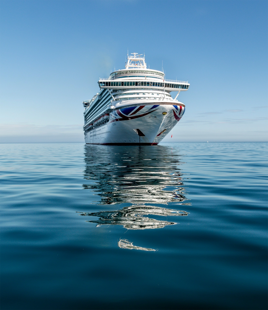 Large white cruise ship sailing across calm blue ocean with its reflection visible in the water under a clear sky. The ship represents the popular cruises from Vancouver Canada that depart along the scenic Pacific coast.
