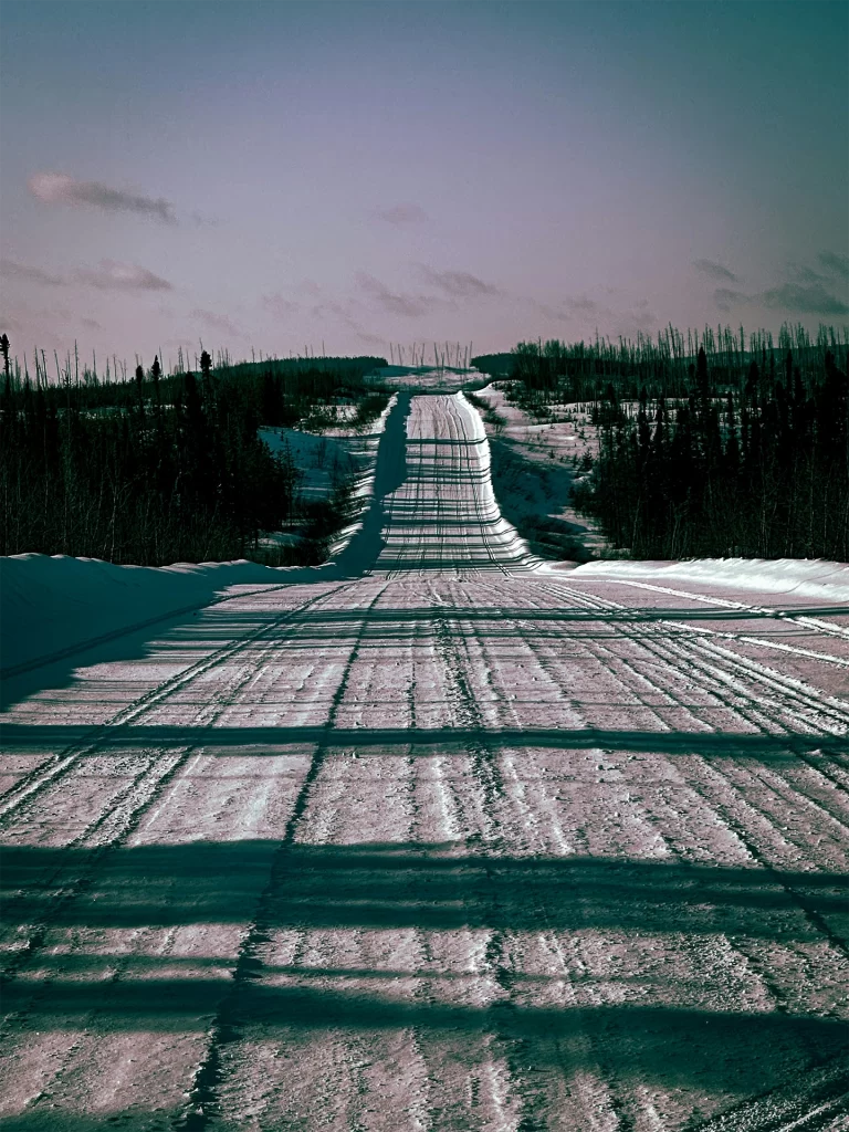 Snow covered road stretching through a remote northern forest with long winter shadows crossing the icy surface under a pale sky. The quiet landscape and open wilderness setting reflect senior travel to Northwest Territories, highlighting scenic winter drives and the peaceful beauty of Canada’s far north.