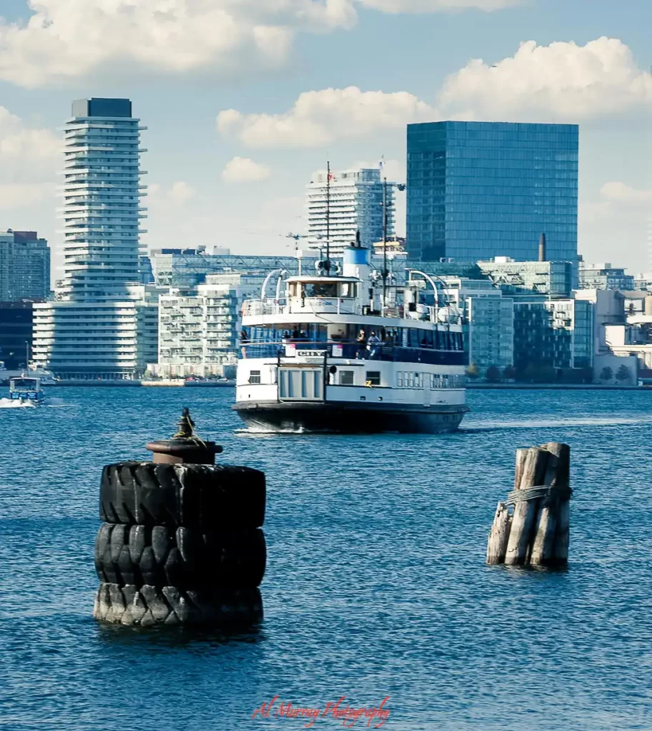 Ferry boat crossing Toronto waterfront with modern condo buildings in the background and wooden posts in the water, showing scenic areas with toronto hotels near public transit and access to ferries and downtown.