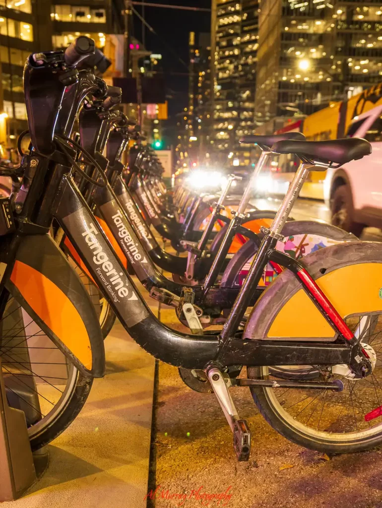 Row of Tangerine branded bike share bicycles lined up on a downtown Toronto street at night with city lights and traffic in the background, showing best areas to stay in toronto without a car with convenient access to cycling and urban transportation.