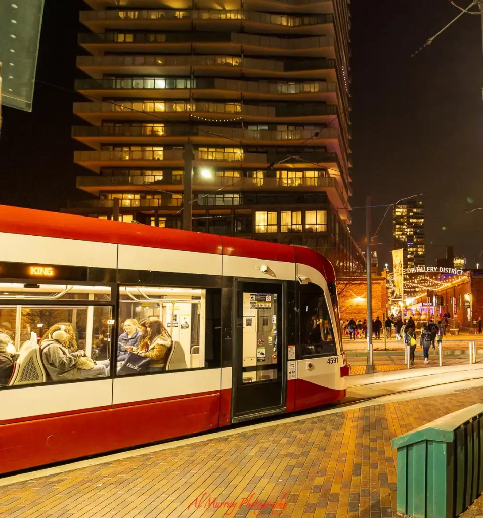 Toronto streetcar stopped at night near the "DISTILLERY DISTRICT" sign with lit buildings and pedestrians walking along a lively street, showing a vibrant area where to stay in Toronto without a car with easy transit access and nightlife nearby.