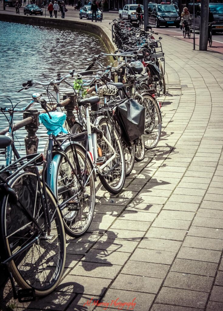 Long row of parked bicycles lines a curved canal side walkway, with rippling water on one side and city life moving quietly in the background. A peaceful urban scene that captures the charm and slow exploration that make scenic getaway ideas feel inviting and memorable.