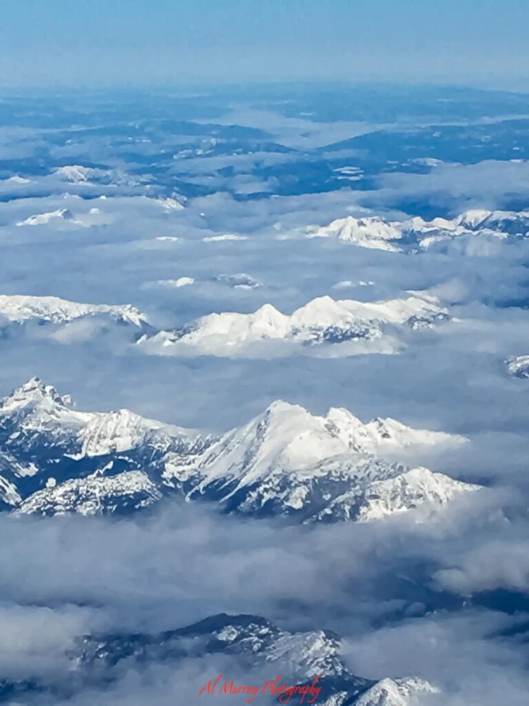 Snow covered mountain peaks rise through layers of soft clouds in a sweeping aerial view, with blue sky fading into the distance above. A peaceful high altitude landscape that inspires scenic getaway ideas focused on quiet beauty, fresh air, and awe inspiring natural views.
