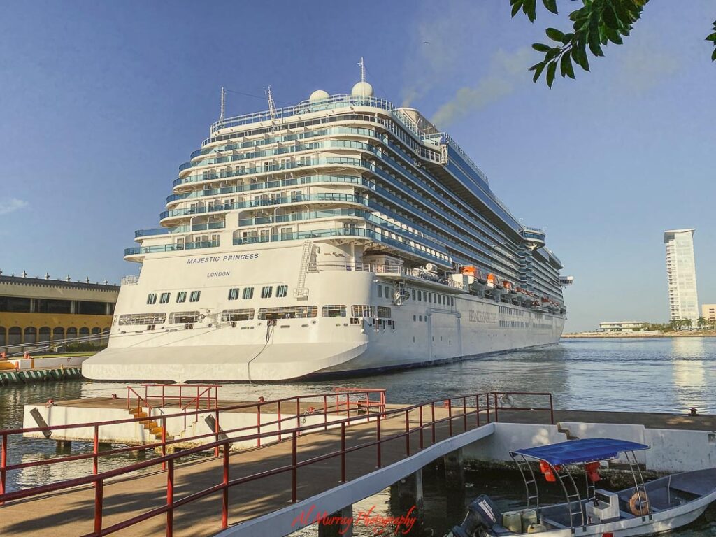 Large white cruise ship docked at a sunny port, towering above the calm water with rows of balconies, lifeboats, and a waterfront skyline in the distance. A clear view of cruise ship size and atmosphere that helps travelers picture the onboard experience when asking if cruising is right for me.