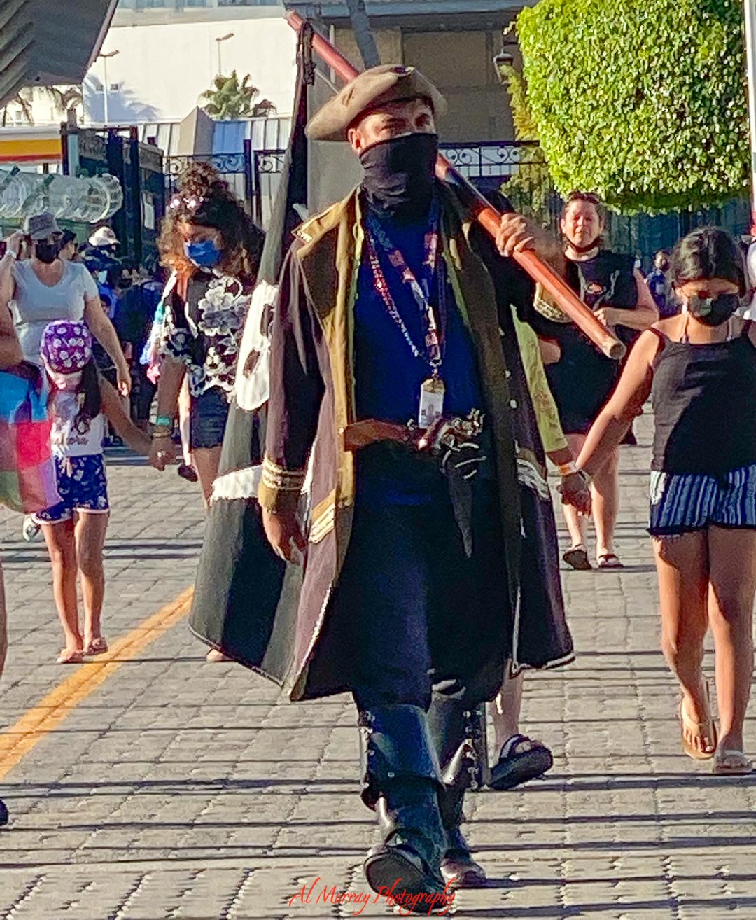 Costumed performer dressed in colonial era clothing walks through a busy cruise port promenade carrying a large flagpole, with travelers strolling on either side in the afternoon sun. A lively shore excursion moment that helps show the culture and atmosphere travelers may consider when deciding if cruising is right for me.