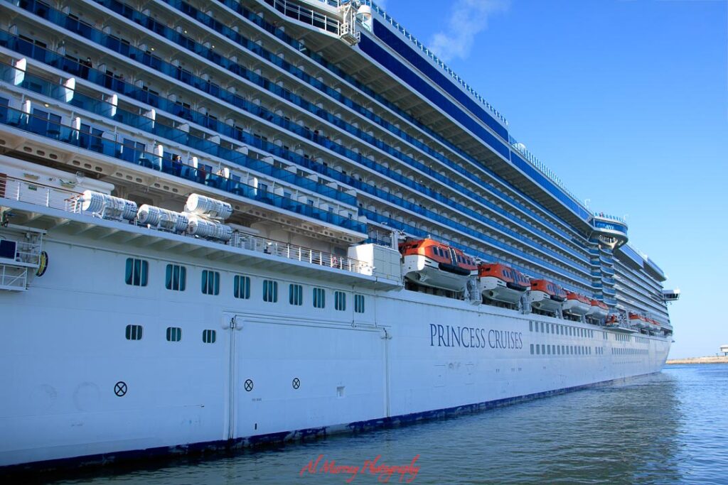 Side view of a large white cruise ship docked in calm water, with long rows of balcony cabins, lifeboats, and spacious upper decks stretching into the distance under a clear blue sky. The ship’s size and variety of onboard spaces help show why cruises are good for multigenerational travellers, offering activities, dining, and room options that can suit different ages and travel styles.