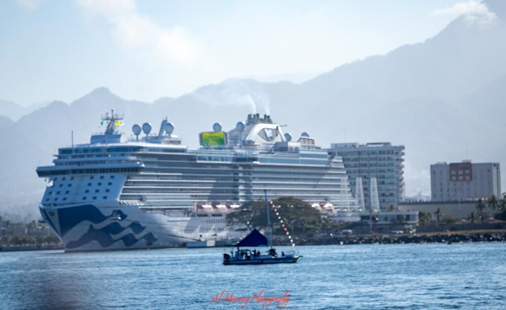 Large cruise ship docked along a calm shoreline with hazy mountains in the background and a small boat crossing the water in the foreground. The contrast between the floating resort and quiet coastal setting helps illustrate what type of traveller enjoys cruises, especially those who like comfort, scenic destinations, and a relaxed way to explore multiple places.