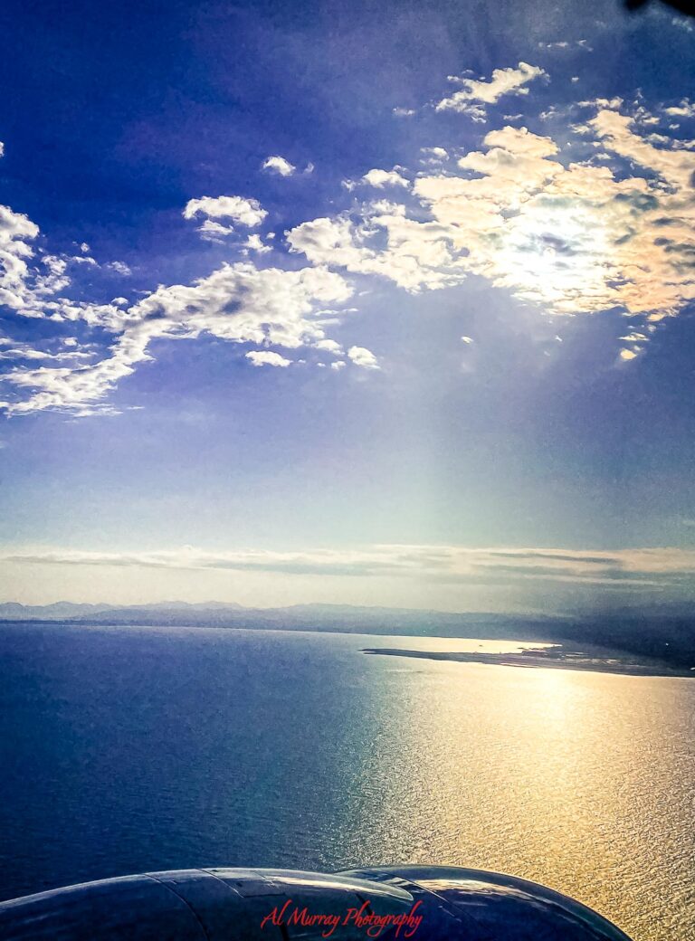 Sunlight breaks through scattered clouds over a calm blue coastline, casting a golden reflection across the water as seen from an airplane window. A peaceful aerial view that captures the quiet beauty of relaxing travel getaways and the sense of escape that comes with them.
