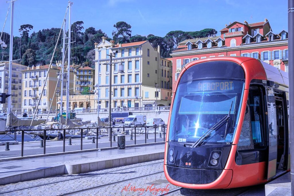 Red modern tram parked along a quiet waterfront promenade, with sailboats, pastel buildings, and tree covered hills in the background under a clear blue sky. A calm coastal city scene that feels ideal for quiet getaway ideas centered on slow travel, scenic views, and peaceful exploration.