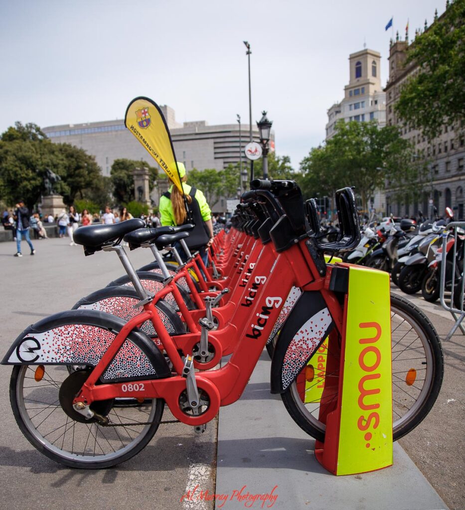 Row of bright red rental bicycles lined up in a city square, with people walking and historic buildings in the background. An easy bike share scene that reflects the slow paced freedom and local exploration often found on relaxing travel getaways.