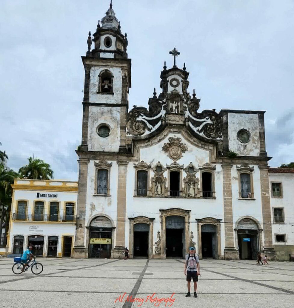 An ornate historic church with twin bell towers stands over a wide stone plaza, with a few visitors walking and cycling across the open square beneath a cloudy sky. The grand architecture and quiet sense of discovery reflect the cultural richness and memorable stops that shape an Atlantic Crossing Cruise Experience.