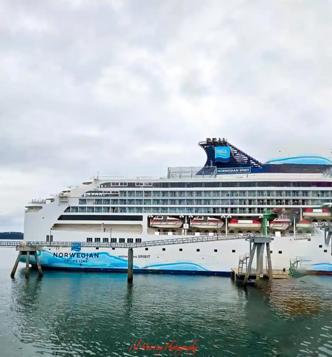 A large white Norwegian cruise ship sits docked beside calm water under a soft gray sky, with rows of balconies, lifeboats, and the bright blue funnel standing out against the muted backdrop. The scale of the vessel and quiet port setting capture the anticipation and comfort that define a Pacific Crossing Cruise Experience.