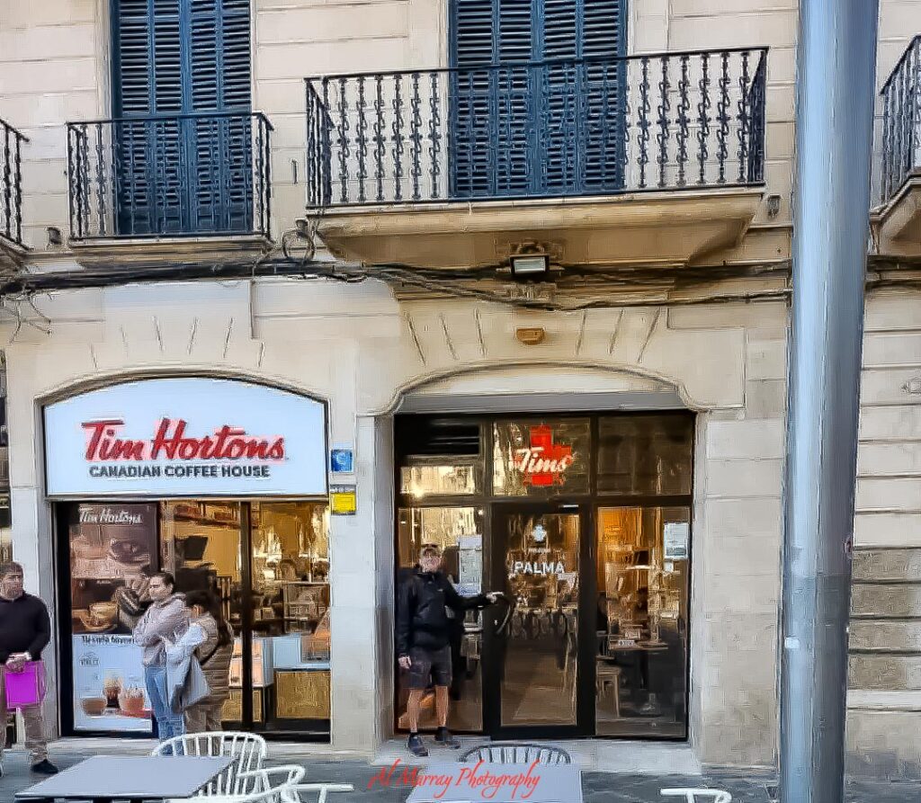 A street level café scene shows a Tim Hortons storefront beneath wrought iron balconies, with pedestrians passing by and outdoor seating in the foreground. This everyday port stop moment highlights the contrast in sea days vs port days cruise travel, where quiet days onboard give way to exploring local streets, coffee shops, and city life ashore.
