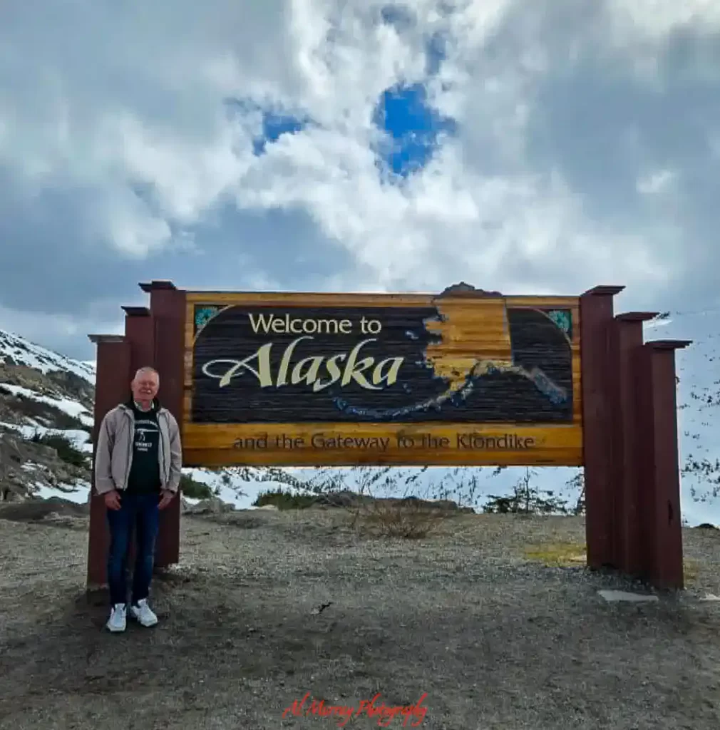 Traveler standing beside a large wooden sign that reads “Welcome to Alaska and the Gateway to the Klondike” with snow covered mountains and dramatic clouds in the background. This scenic stop captures the rugged landscapes and memorable destinations often included in a Pacific Crossing Cruise Experience.