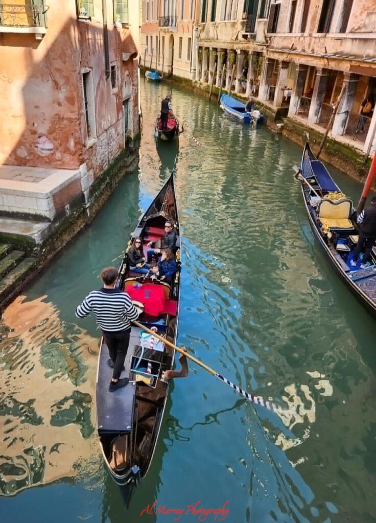 Several gondolas glide through a narrow Venetian canal bordered by weathered pastel buildings, with passengers seated as gondoliers steer each boat along the green waterway. This relaxed sightseeing moment captures the charm, comfort, and cultural exploration often highlighted in a mature traveller cruise review.